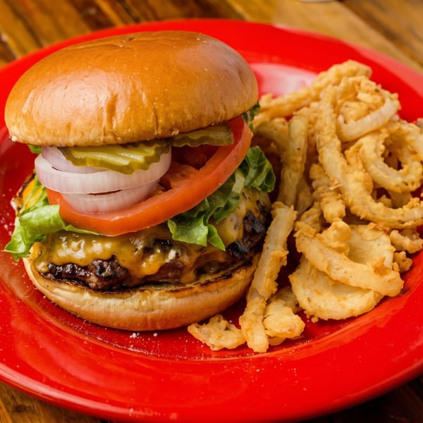 Cheeseburger with lettuce, tomato, onion, and pickles, served with crispy onion rings on a red plate.