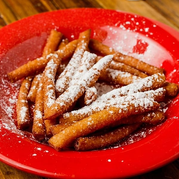 A red plate filled with golden-brown churros dusted with powdered sugar on a wooden table.