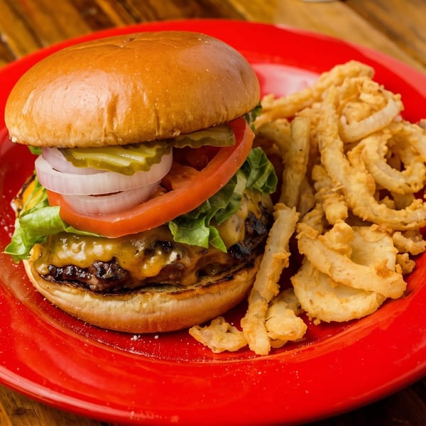 Cheeseburger with lettuce, tomato, onion, and pickles, served with crispy onion rings on a red plate.